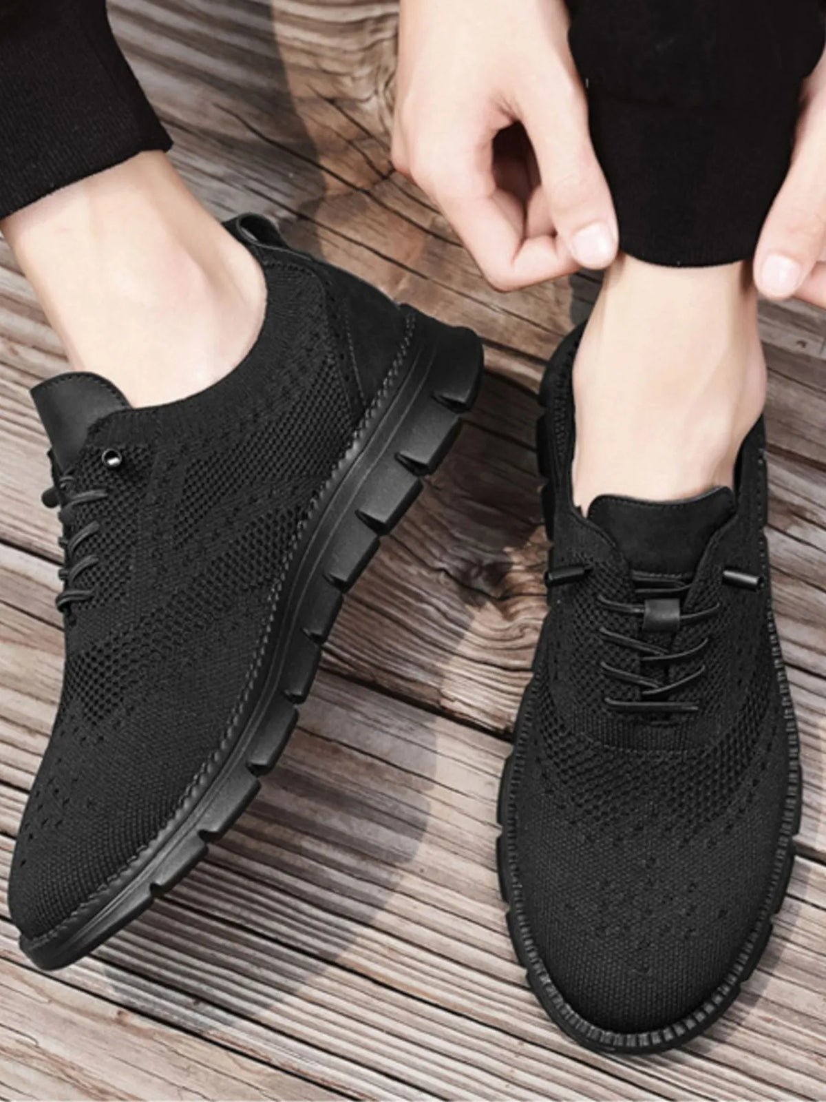 Man lacing black knit mesh lace-up sneakers with black rubber soles on a wooden floor