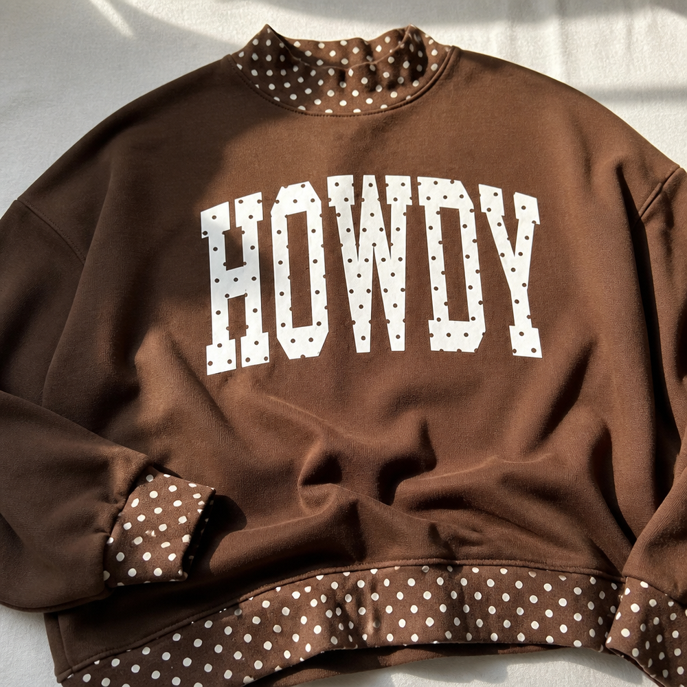 A brown crewneck sweatshirt with 'HOWDY' printed in white polka-dot block letters across the chest. The inner collar, cuffs, and hem are also brown with a white polka-dot pattern, laid flat on a light fabric background.