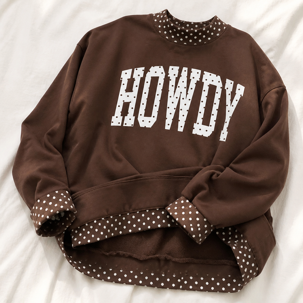 A brown crewneck sweatshirt with 'HOWDY' printed in white block letters on the chest, featuring white polka-dot trim on the collar, cuffs, and hem, laid flat on a white fabric background.
